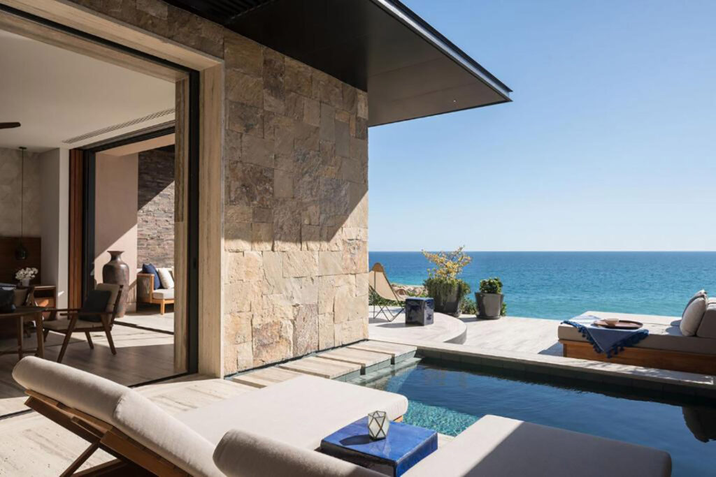 Exterior shot of luxury hotel with limestone outside and small pool with deck chairs overlooking blue ocean and clear skyline
